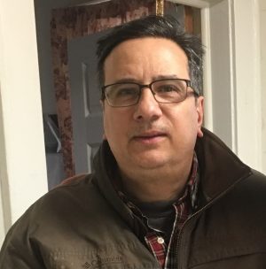 A middle-aged man with glasses and dark hair, wearing a brown jacket and plaid shirt, stands indoors in front of a doorway.
