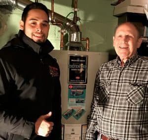 Two men stand indoors next to a water heater; one gives a thumbs up while both smile at the camera.