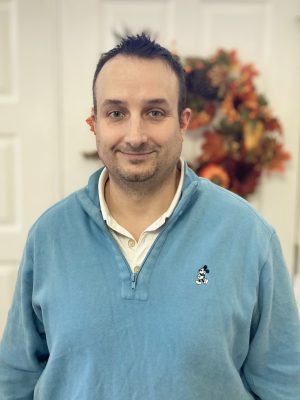 A man wearing a light blue Mickey Mouse pullover stands indoors in front of a white door with a fall-themed wreath in the background.