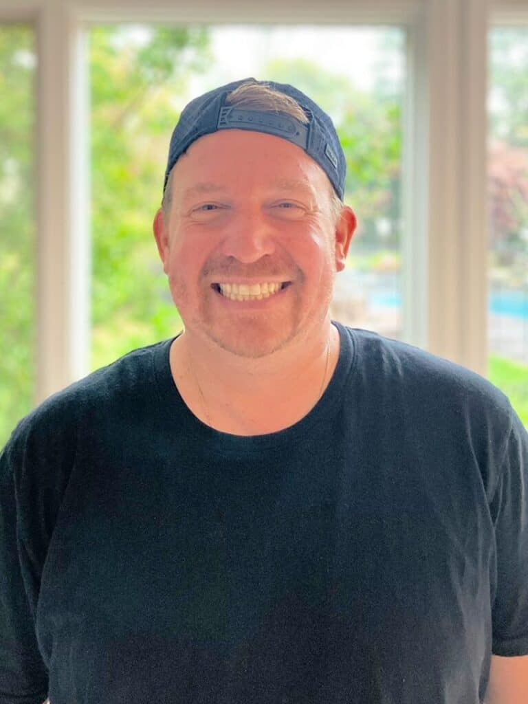 A man wearing a black t-shirt and a backwards cap is smiling while standing indoors in front of large windows with a leafy background.