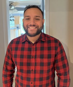 A man with short dark hair and a beard is smiling, wearing a red and black checkered shirt, standing indoors with light coming from behind.