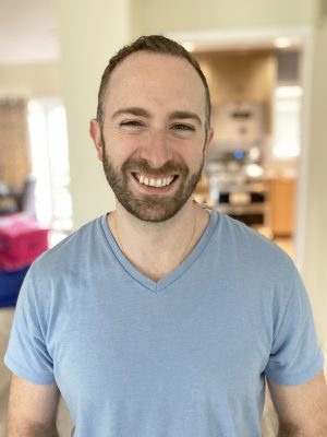 A man with short hair and a beard, wearing a light blue V-neck T-shirt, smiles while standing indoors in a brightly lit room.