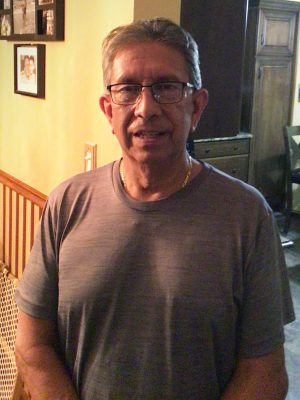 An older man with glasses and short gray hair, wearing a gray t-shirt, stands indoors near a wooden railing and framed photos on the wall.