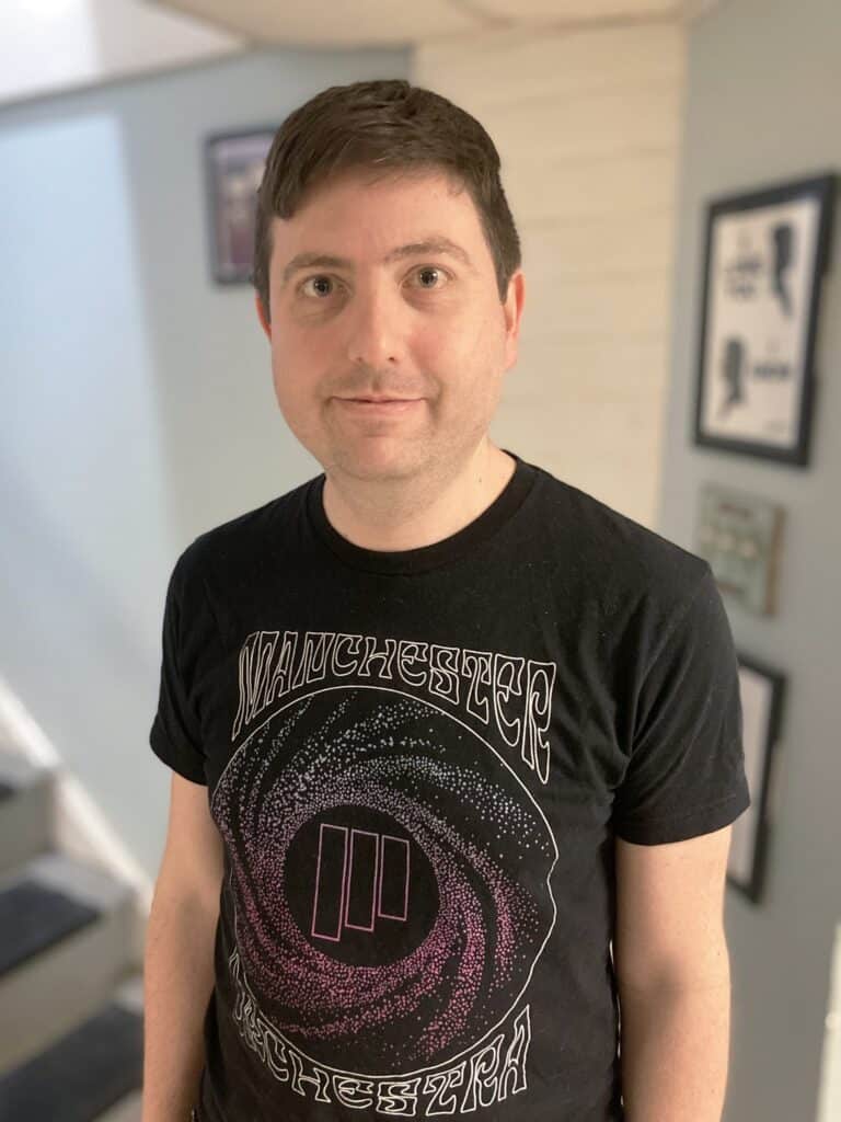 A man with short brown hair stands indoors wearing a black Manchester Orchestra t-shirt; framed pictures hang on the light blue wall behind him.