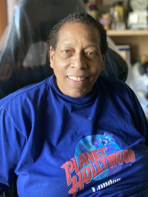 A person wearing a blue Planet Hollywood London t-shirt sits indoors and smiles at the camera.