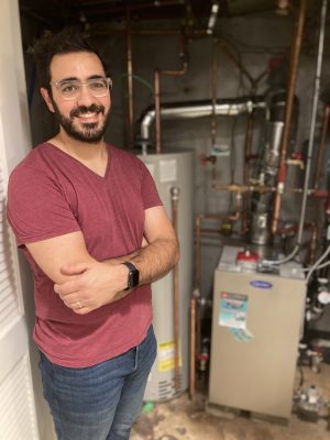 A man with glasses and a beard stands with arms crossed in front of a water heater and furnace in a utility room.