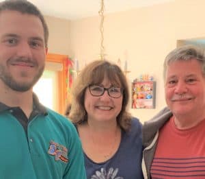 Three people stand close together indoors, smiling at the camera. The background includes a window, decorations, and a wall shelf with items on it.
