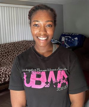 A woman smiling indoors, wearing a black T-shirt with pink and white lettering reading "Undergraduate Business Women's Association 2012-2013." A bed and a couch are visible in the background.