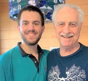 Two men, one younger and one older, stand close together indoors, smiling at the camera. A stained glass light fixture hangs above them.
