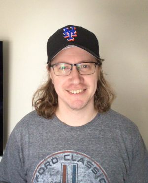 A person with long hair and glasses smiles at the camera, wearing a black baseball cap and a gray Ford Classic t-shirt.