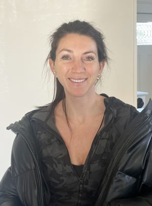 A woman with long dark hair wearing a black zip-up jacket and a camouflage-patterned top smiles at the camera indoors.