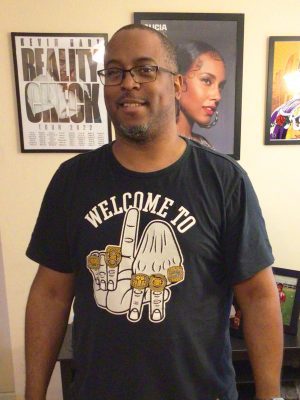 A man wearing glasses and a "Welcome To" t-shirt stands indoors in front of framed posters on a wall.