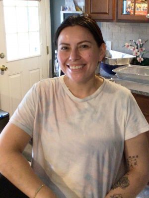 A woman with dark hair, wearing a light-colored t-shirt, sits and smiles in a kitchen with wooden cabinets and a door in the background.