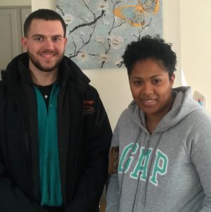 Two people standing indoors and facing the camera, one wearing a black jacket and the other wearing a grey GAP hoodie. A floral painting is visible on the wall behind them.