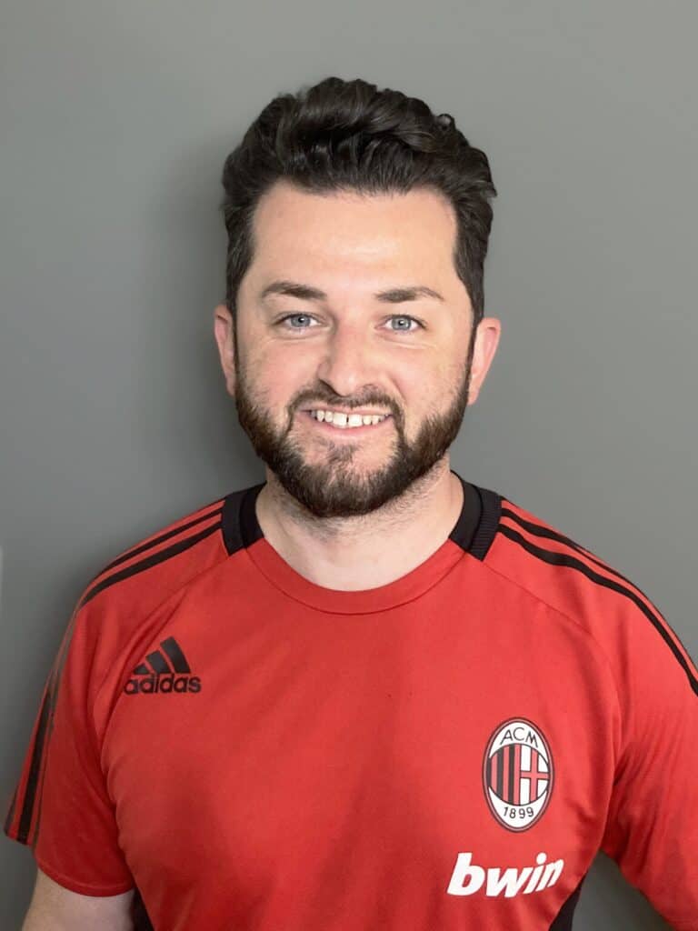 Man with short dark hair and beard wearing a red AC Milan soccer jersey, standing in front of a plain gray background and smiling at the camera.
