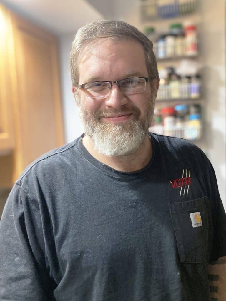 A man with glasses and a gray beard stands indoors wearing a black t-shirt. Shelves with spice jars are visible in the background.