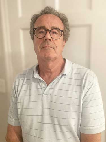 An older man with gray hair and glasses wearing a light-colored, short-sleeve, striped polo shirt stands in front of a white door.