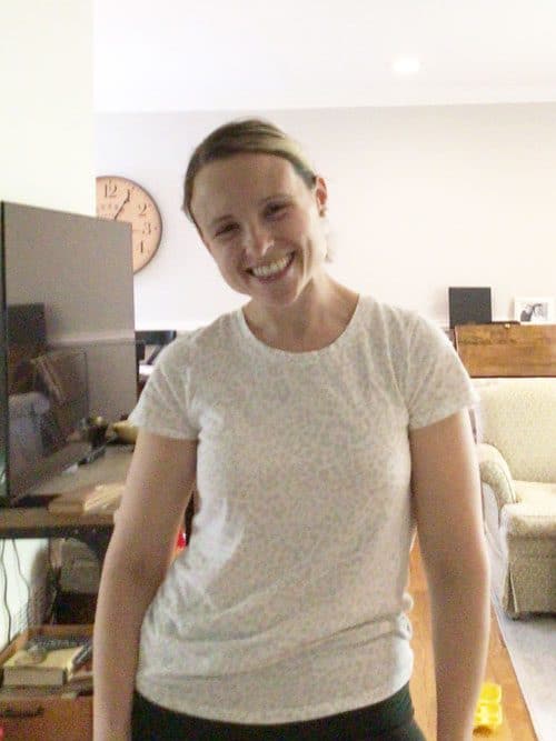A woman standing and smiling in a living room, wearing a white patterned t-shirt. There is a TV, a clock on the wall, and some furniture in the background.