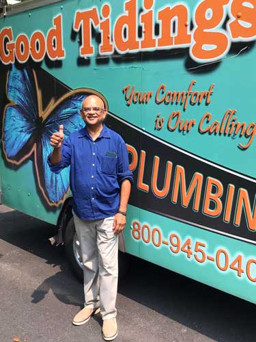 A man in a blue shirt and beige pants stands in front of a plumbing service van, smiling and giving a thumbs-up.