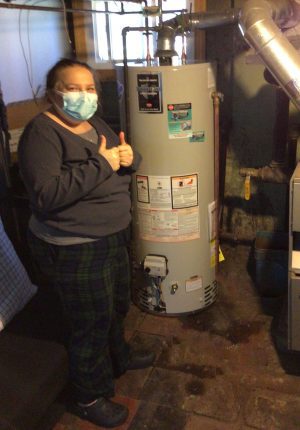 A person wearing a mask gives a thumbs up while standing next to a water heater in a basement.