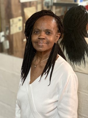 A woman with long braids and a white sweater stands indoors, smiling at the camera with a blurred background and mirror behind her.