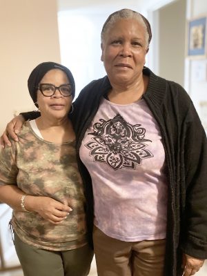 Two women stand side by side indoors, one wearing glasses and a camouflage shirt, the other in a purple shirt with a black cardigan.
