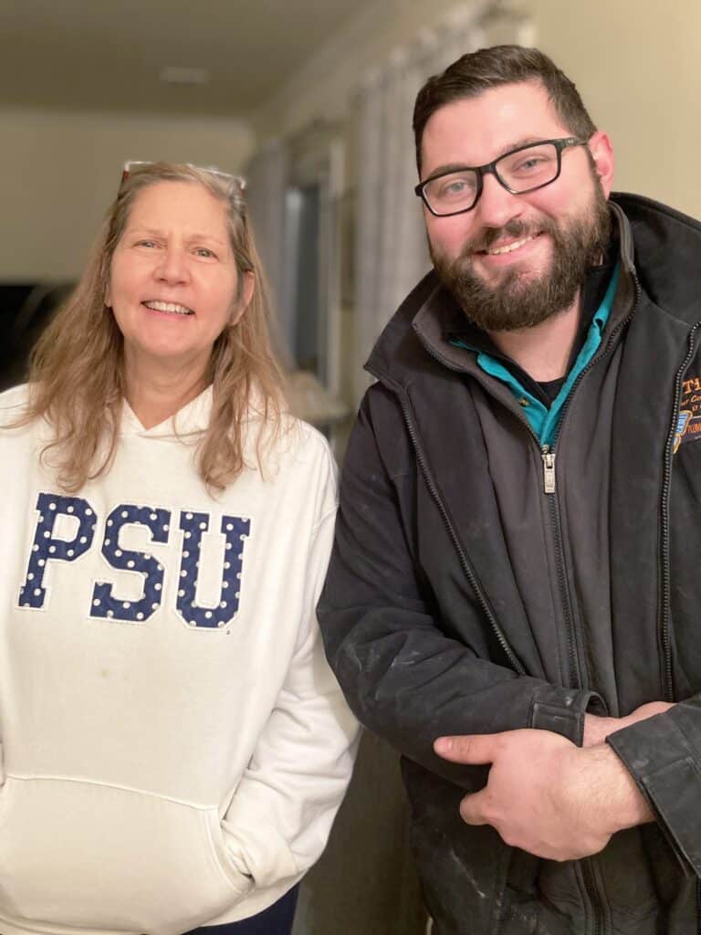Two people stand indoors smiling at the camera. The person on the left wears a white PSU sweatshirt; the person on the right wears glasses and a dark jacket.