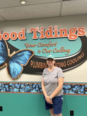 A person in casual clothes stands in front of a wall with a "Good Tidings" plumbing, heating, and cooling sign featuring a large blue butterfly.