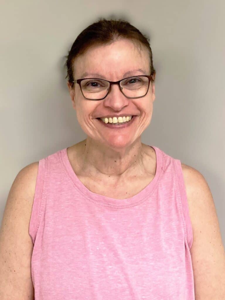 A woman with glasses and brown hair pulled back is smiling at the camera. She is wearing a sleeveless pink top and standing against a plain beige background.