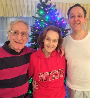 Three people stand smiling in front of a decorated Christmas tree; the woman in the center wears a red Rutgers sweatshirt.