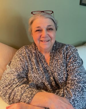 An older woman with gray hair and glasses on her head sits on a couch, smiling at the camera, wearing a patterned top.