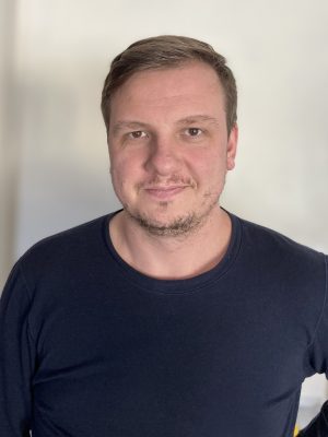 A man with short brown hair and a trimmed beard is standing indoors, facing the camera, wearing a dark long-sleeve shirt against a plain light background.