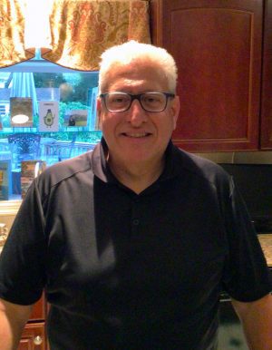A man with short gray hair and glasses stands in a kitchen, smiling at the camera. Brown cabinets and a window with a curtain are visible in the background.