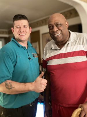 Two men stand indoors smiling at the camera; one in a teal polo shirt gives a thumbs up, while the other wears a red and gray shirt.