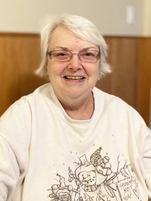 An older woman with short white hair and glasses smiles at the camera, wearing a white sweatshirt with a snowman graphic.