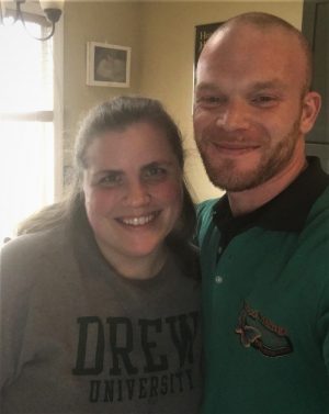 Two people smiling indoors; the woman is wearing a Drew University shirt and the man is wearing a green and black collared shirt.