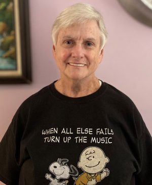 Older adult with short gray hair smiles at the camera, wearing a black T-shirt with Snoopy and Charlie Brown and the words "When all else fails turn up the music.