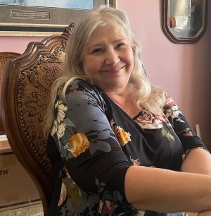 An older woman with long gray hair sits on a carved wooden chair, smiling, wearing a dark floral top, in a room with pink walls and framed art.