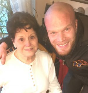 An older woman sits and smiles at the camera while a younger man stands next to her, leaning in with his arm around her shoulder.