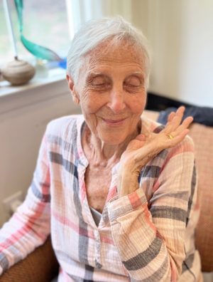 Elderly woman with short white hair sits in a chair, eyes closed, smiling softly, and resting her hand near her face. She wears a plaid shirt.
