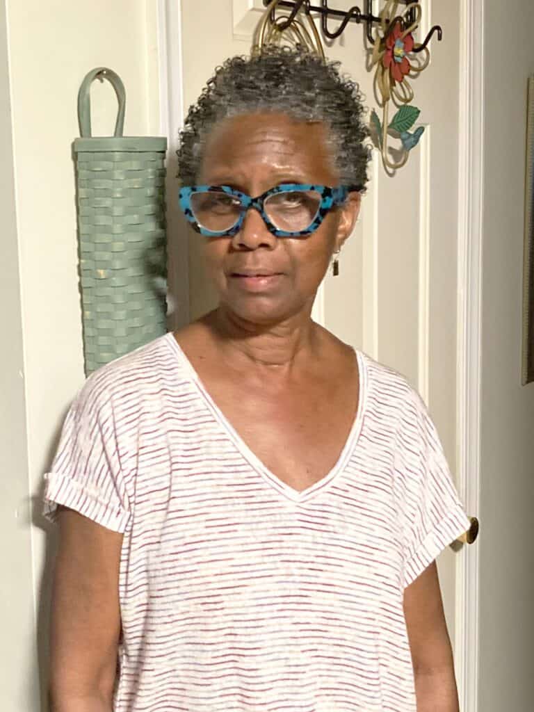 An older woman with short curly hair and blue patterned glasses stands indoors, wearing a white striped shirt, near a green woven basket hanging on the wall.