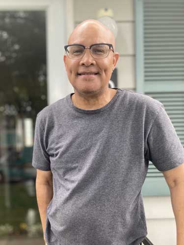 A man wearing glasses and a gray t-shirt stands outside in front of a house with light-colored siding and blue shutters, smiling at the camera.