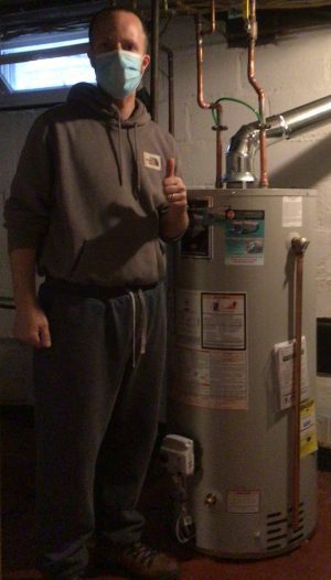 A person wearing a face mask and hoodie stands next to a water heater in a basement, giving a thumbs-up gesture.