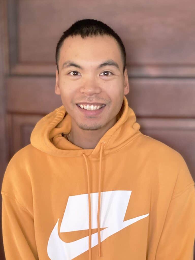 A person wearing an orange hoodie with a white abstract logo stands in front of a wooden background, smiling at the camera.