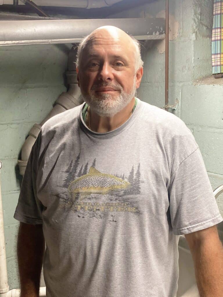 A middle-aged man with a gray beard stands in a basement, wearing a gray T-shirt with a fish and forest design. Exposed pipes and cinder block walls are visible in the background.
