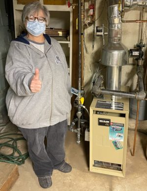 A person wearing a mask and gray sweatshirt gives a thumbs up while standing next to a beige furnace in a basement.