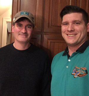 Two men stand indoors in front of wooden cabinets, one wearing a hat and dark sweater, the other in a teal polo shirt with an embroidered logo, both smiling at the camera.