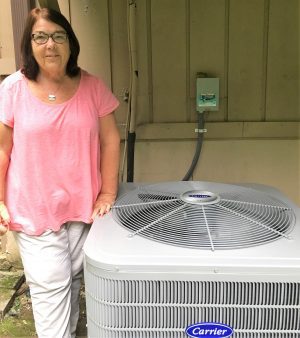 A woman in a pink shirt stands next to a large outdoor Carrier air conditioning unit beside a house.