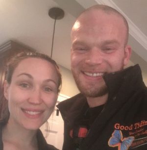 Two people are smiling indoors; the man is wearing a black jacket with a butterfly and the words "Good Tidings" on it.