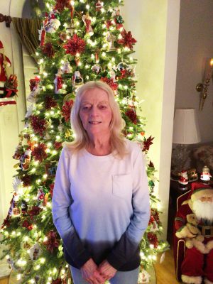 A woman stands in front of a decorated and lit Christmas tree in a living room, with a Santa Claus figure and lamp visible to the side.